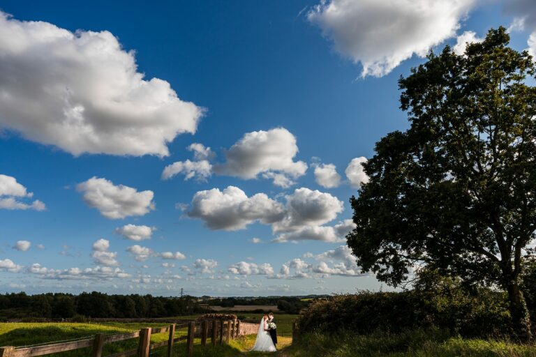 A couple dressed in wedding attire poses under a vast, clear blue sky dotted with fluffy white clouds. They stand beside a wooden fence in a scenic countryside landscape with lush green fields, a tall tree on the right, and distant trees on the horizon. Image by Documentary Wedding Photographer Lewis Bishop Photography.