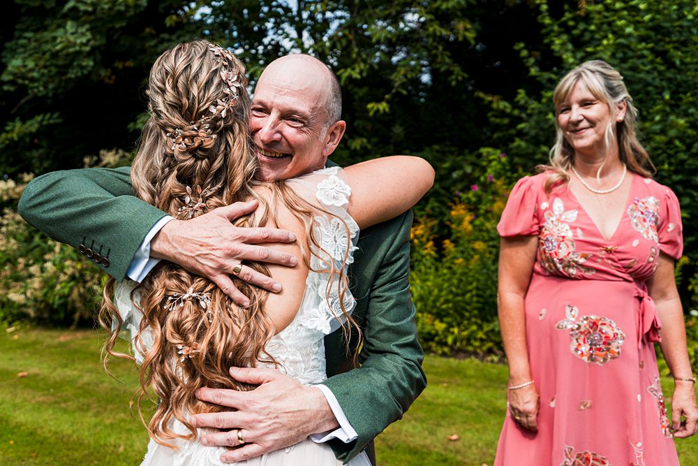 Bride hugging man with woman in pink dress smiling.
