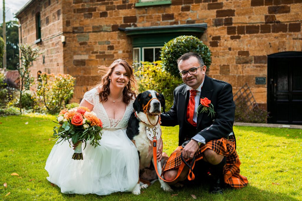 Bride and groom with dog outside stone building.