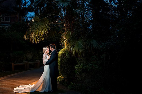 Bride and groom embrace under illuminated tree.