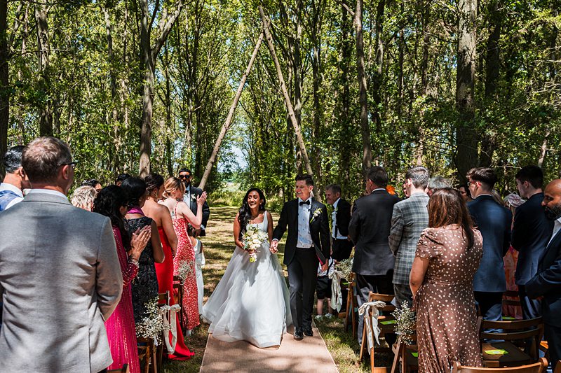 Bride and groom walk down forest aisle.