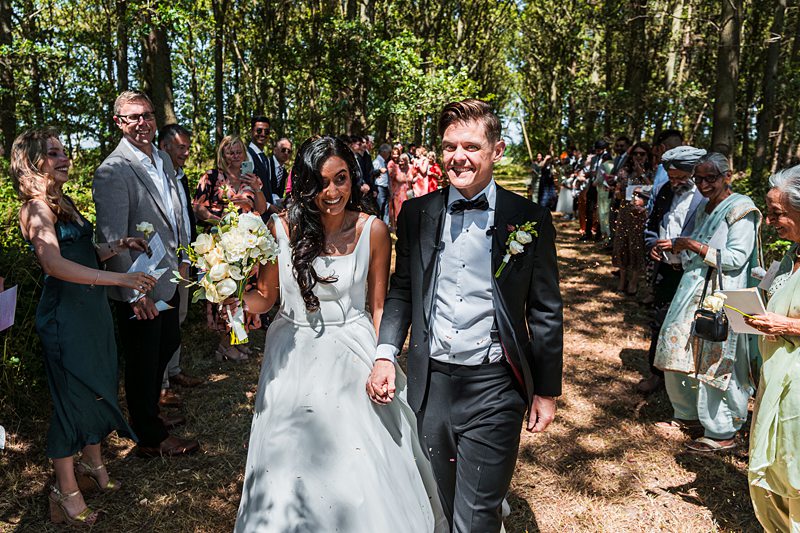 Bride and groom walk down outdoor aisle, smiling.