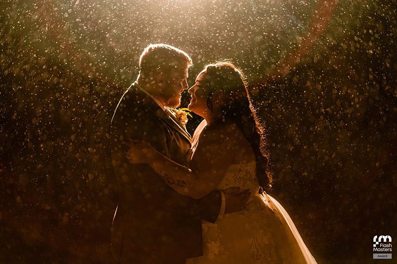 Bride and groom embrace romantically in rain.