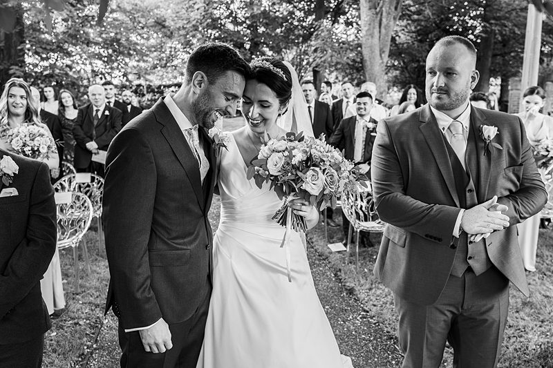 Bride and groom smiling during outdoor wedding ceremony.