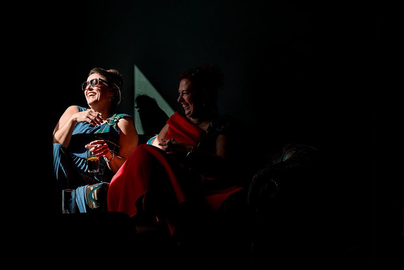 Two women laughing, seated in dim light.