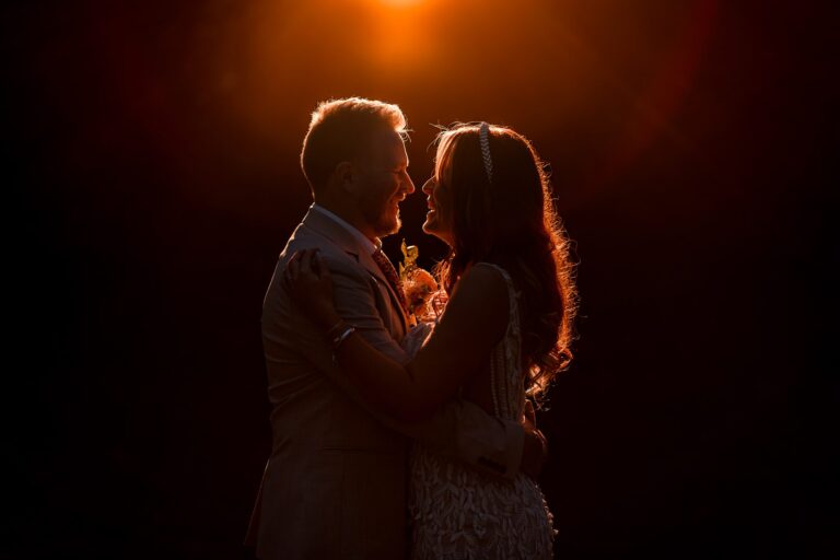 Couple embraces at sunset, silhouetted against orange sky.
