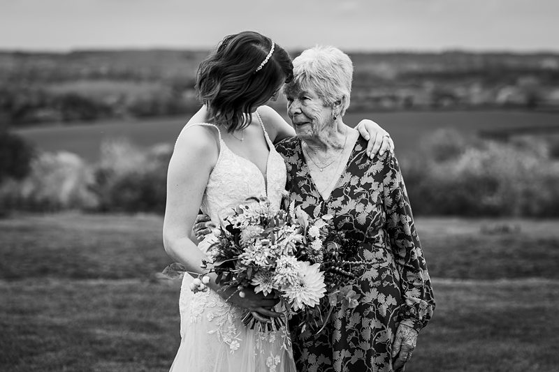 Bride embraces elderly woman in countryside setting.