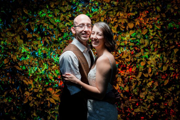 Smiling couple embraces in front of colourful leaves.