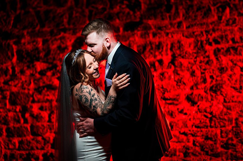 Bride and groom embrace against red brick wall.