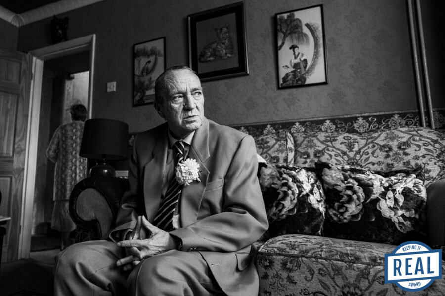 Elderly man in suit sitting on patterned sofa.